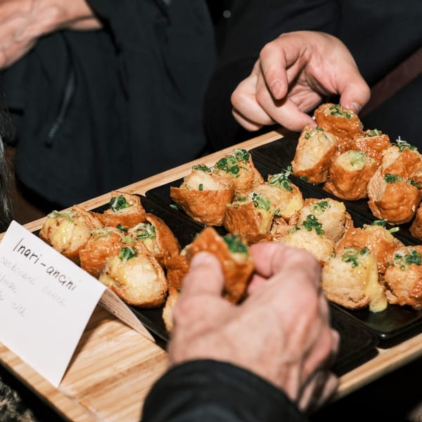 Hands reaching for Nem's Japandi Bistro's Inari-ancini, Japanese fried rice balls, from a wooden tray.