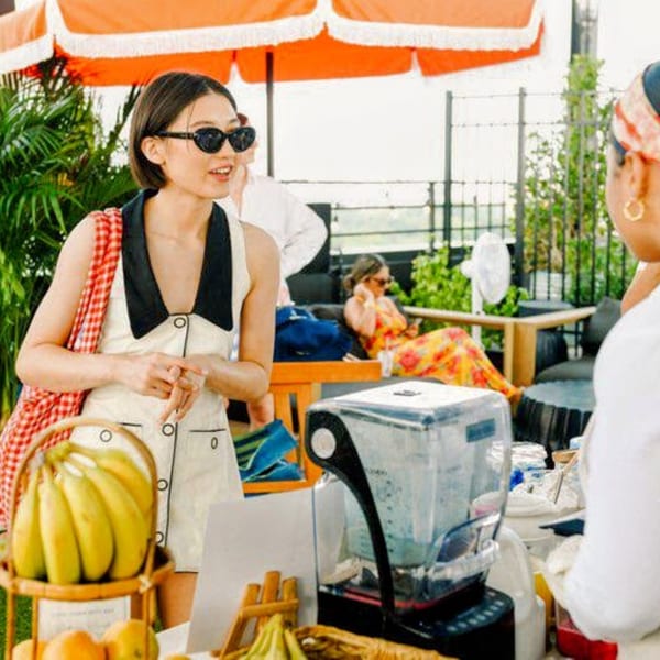 A woman in sunglasses and a white dress stands at an outdoor bar with bananas and a blender.