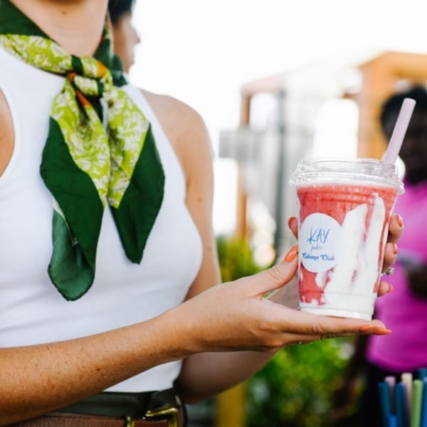 A person in a white top and green scarf holds a strawberry and cream smoothie with a pink straw.