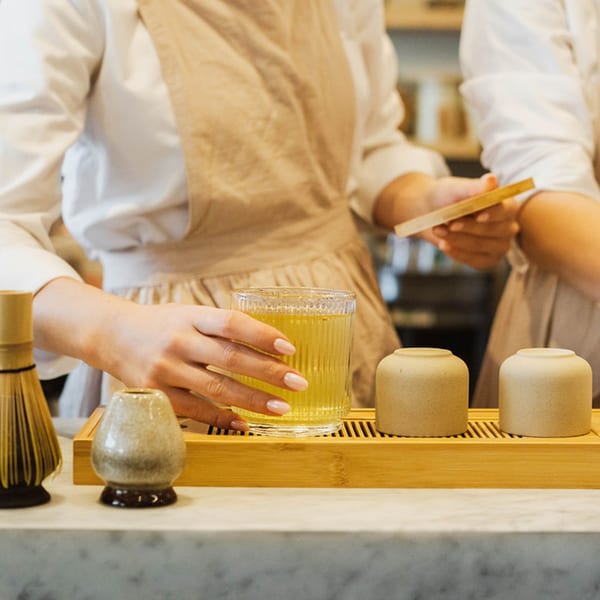 A server in an apron places a glass of green tea on a wooden tray with Japanese tea ceremony items.