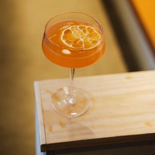 An orange cocktail with a dried orange slice garnish sits on a light wooden surface.