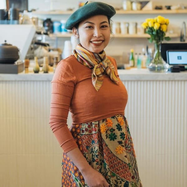 Smiling woman in a dark beret, orange top, and patterned skirt stands in Nem's Japandi Bistro.