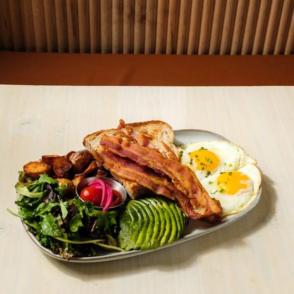 A breakfast plate with bacon, fried eggs, avocado, roasted potatoes, and a side salad.