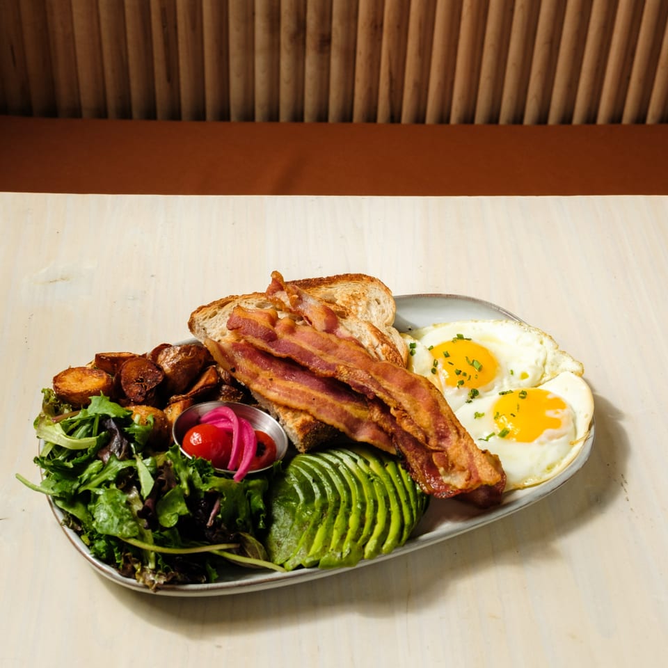 A breakfast plate with bacon, fried eggs, avocado, roasted potatoes, and a side salad.