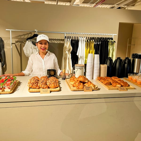 A smiling woman in a white cap stands behind a counter with croissants, parfaits, and coffee.