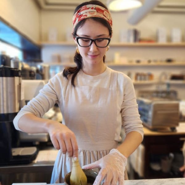 A Nem's Japandi Bistro employee in glasses and apron prepares a drink with a whisk.