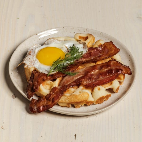 Waffles topped with crispy bacon, a fried egg with a bright yellow yolk, and fresh dill.